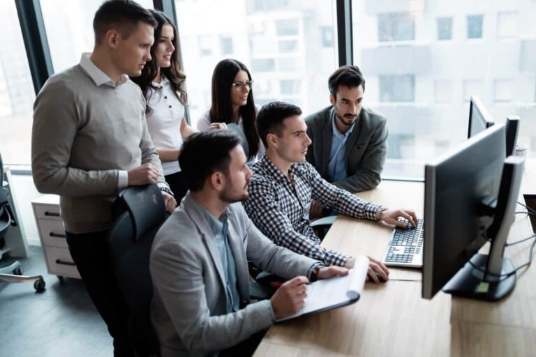 A group of young professionals gathered around a desk, looking at a computer monitor together in an office setting.