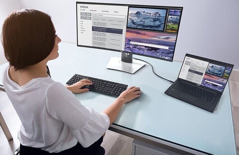 Persona usando una estación de trabajo de dos pantallas: un monitor grande y una computadora portátil conectada en un escritorio de vidrio, que muestran correos electrónicos y fotos uno al lado del otro.