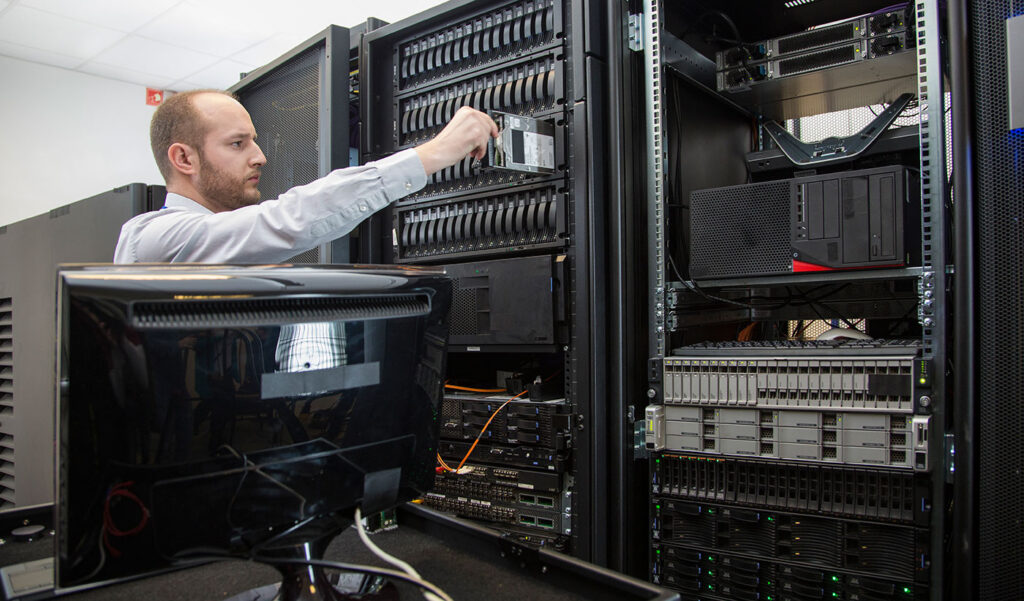 IT-Techniker in einem Rechenzentrum schiebt eine Festplatte in ein Server-Rack zwischen mehreren gestapelten Servern und Netzwerkgeräten ein.